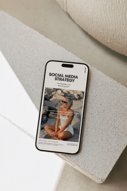 A smartphone lying on a light stone surface, displaying a social media strategy post with a photo of a woman sitting outdoors. The phone is shown in a styled flat-lay setting with neutral stone blocks and a cream fabric surface.